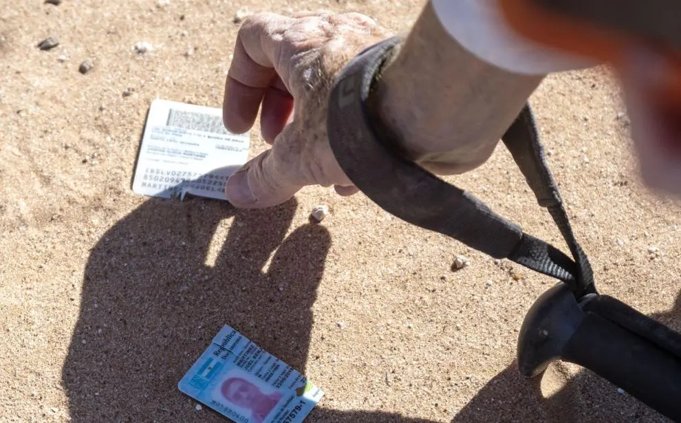 A gloved hand reaches toward personal identification cards lying partially buried in the desert sand, next to the strap of a backpack or field gear.