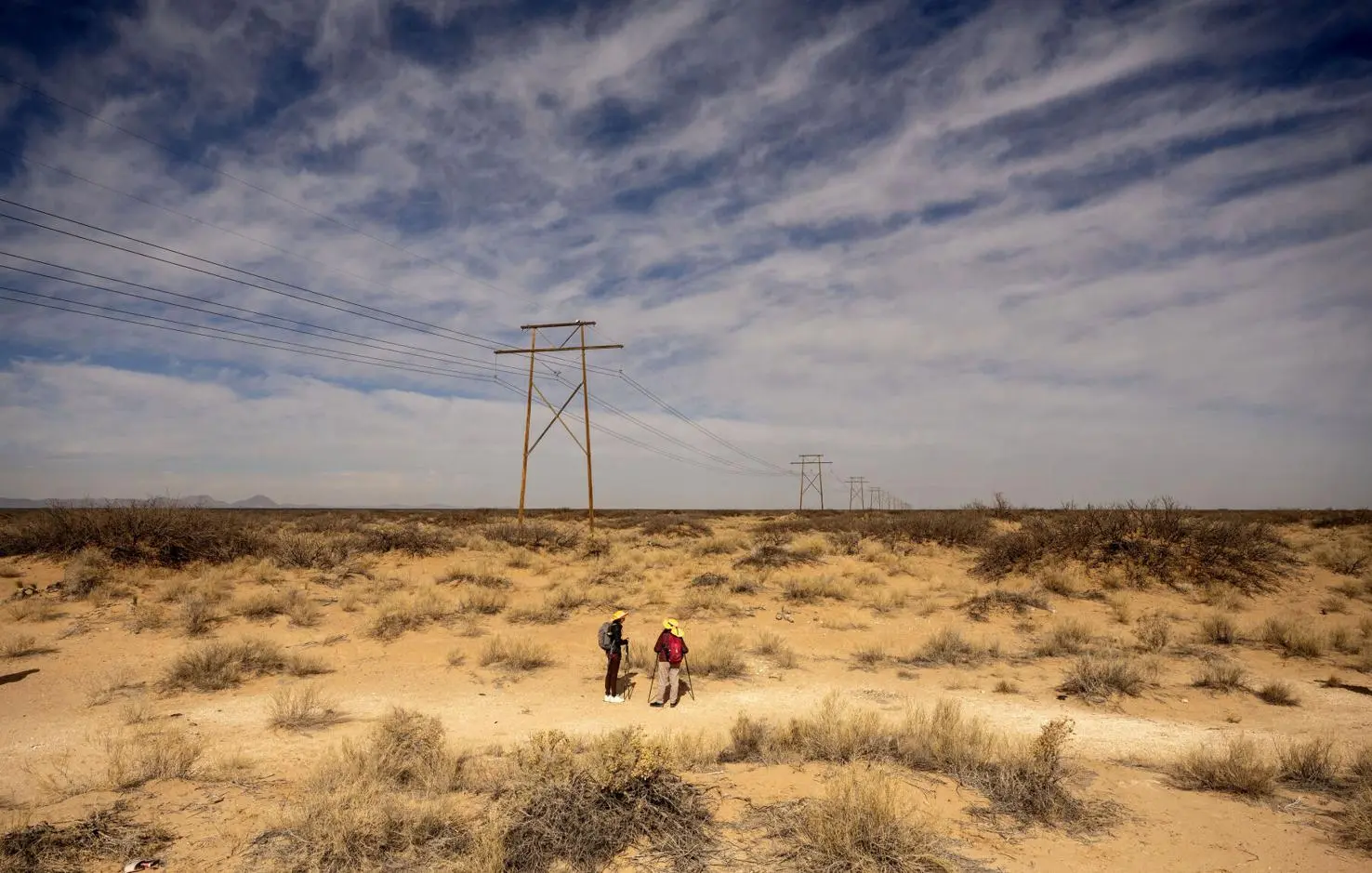 Two people walk across an expansive desert landscape beneath power lines, surrounded by sparse shrubs and dry sand under a wide, cloud-streaked sky.