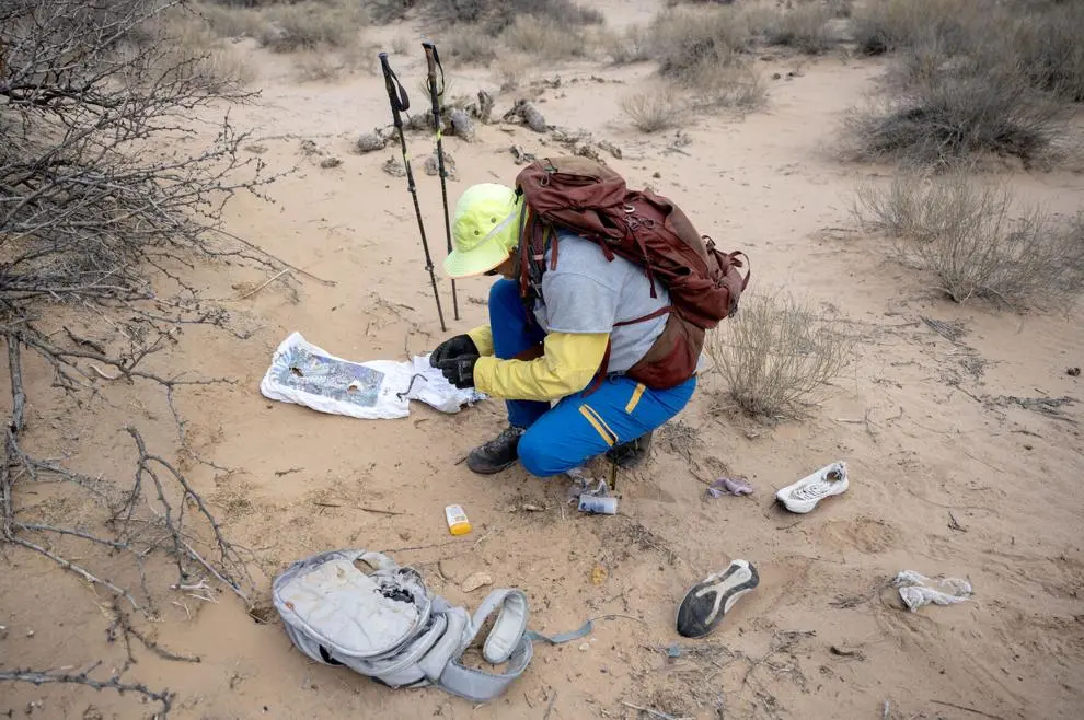 A humanitarian aid volunteer kneels in the desert examining personal items found at a remote site, including shoes, a backpack and scattered belongings, with walking poles nearby.