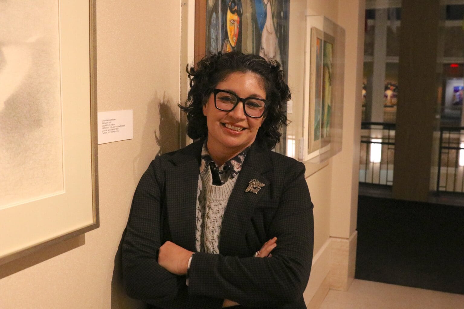 State Rep. Sarah Silva stands indoors in a gallery space with framed artwork on the walls behind her, smiling with her arms crossed.