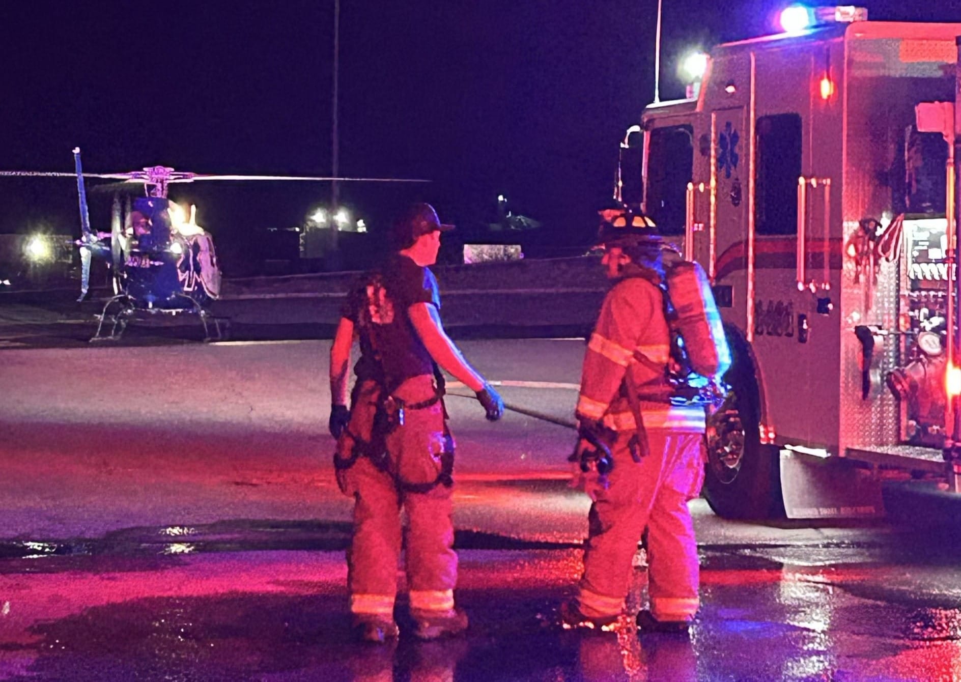 Emergency responders stand beside a fire truck while a helicopter sits nearby during a nighttime response scene.