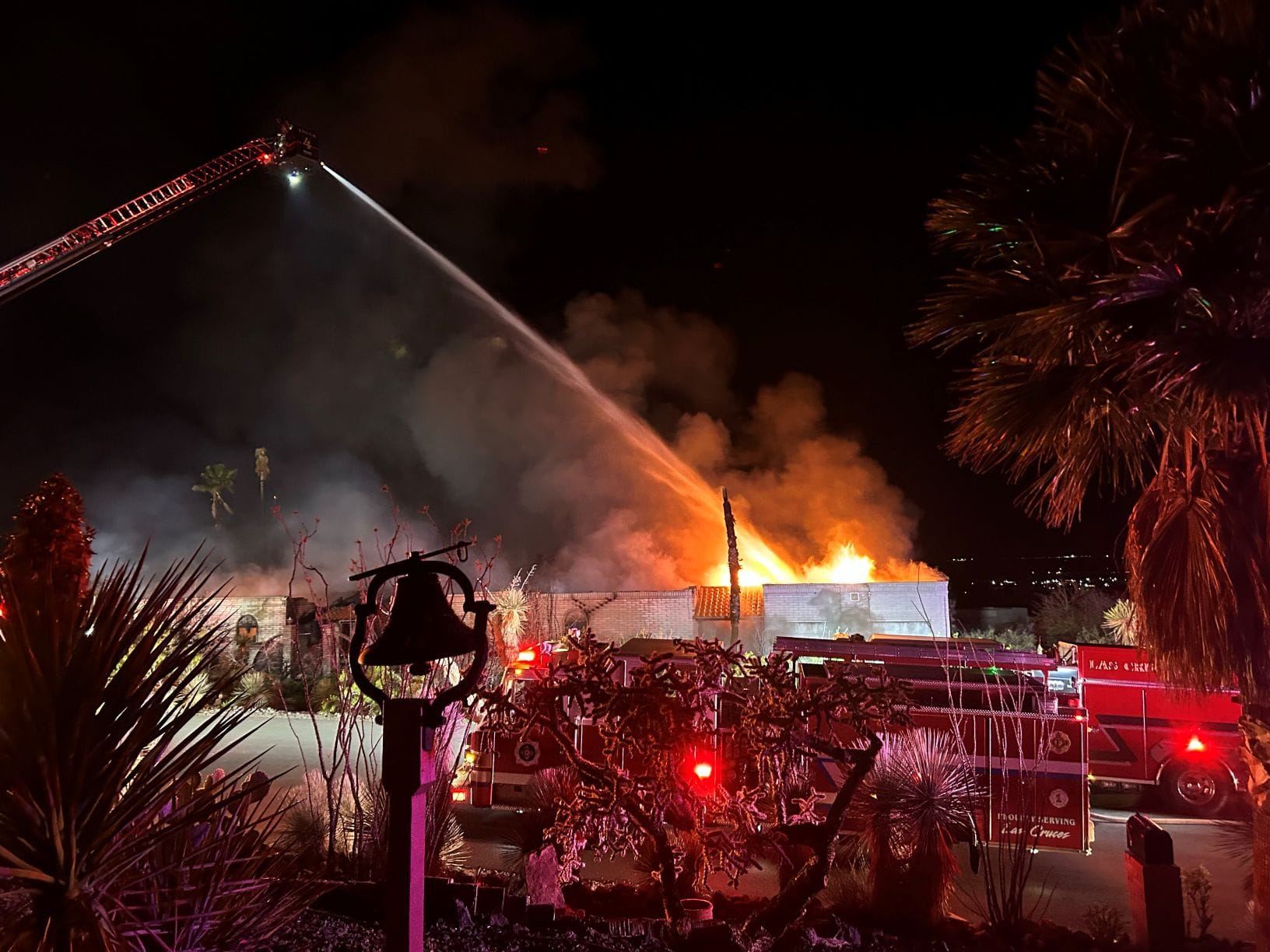 Firefighters spray water from a ladder truck onto a house fire at night, with flames and heavy smoke rising from the structure in the Las Alturas area.