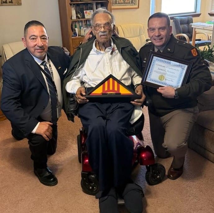 James C. Flowers sits at home in Las Cruces holding a folded flag as two New Mexico veterans officials present him with a certificate and recognition award.