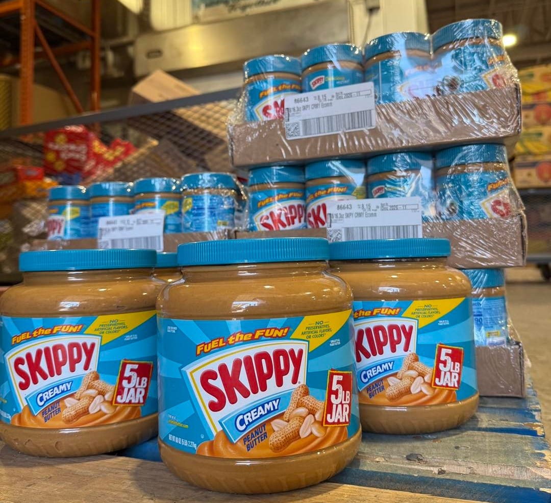 Large jars of Skippy peanut butter displayed in front of stacked cases of peanut butter at a food distribution warehouse.
