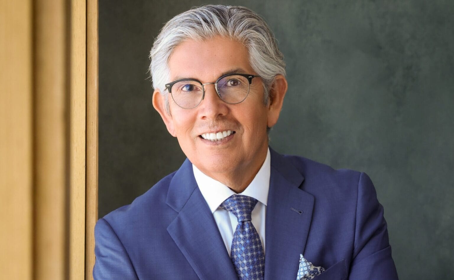 Portrait of Duke Rodriguez wearing a blue suit and glasses, smiling at the camera