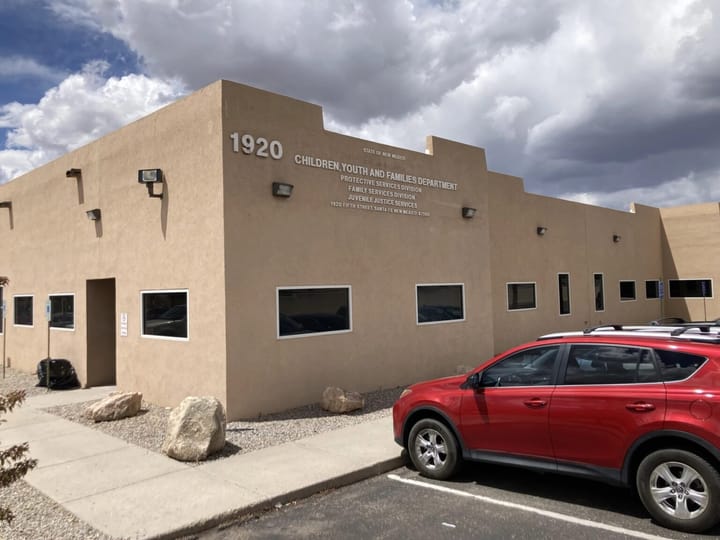 The New Mexico Children, Youth and Families Department building in Santa Fe, with a red SUV parked outside under cloudy skies.