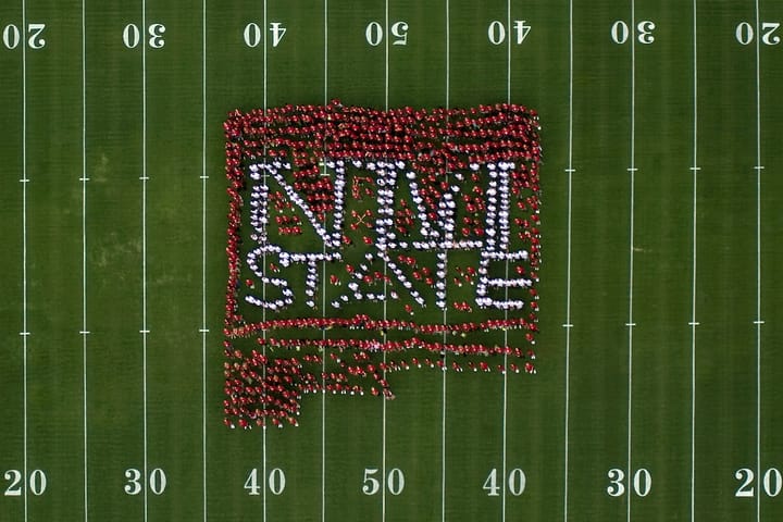 Aerial view of hundreds of New Mexico State University students standing on a football field, arranged to form the NMSU logo and the words “NM State.”