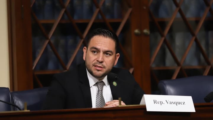 A man in a suit and tie sits at a congressional hearing table speaking into a microphone, with a nameplate reading “Rep. Vasquez” in front of him.