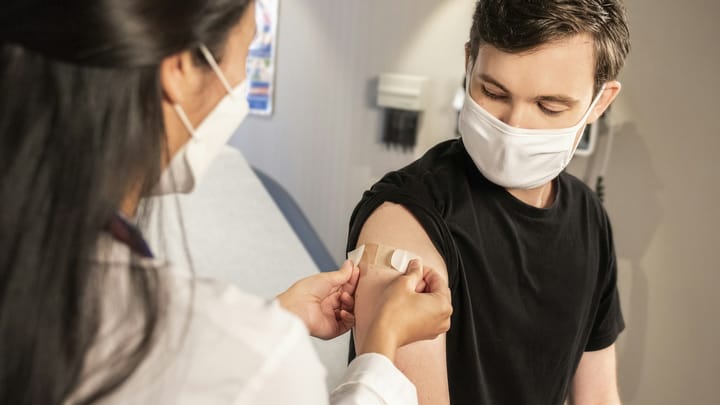 A masked health care worker applies a bandage to a patient’s upper arm after administering a vaccination.