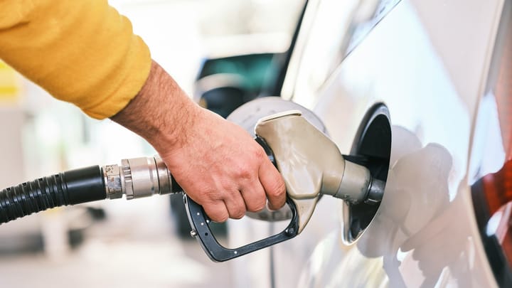 A person wearing a yellow sleeve refuels a car, holding a gas pump nozzle inserted into the vehicle’s tank at a station.