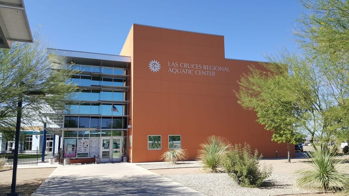 Exterior of the Las Cruces Regional Aquatic Center on East Hadley Avenue, showing its orange and glass facade under a clear blue sky with desert landscaping in front.