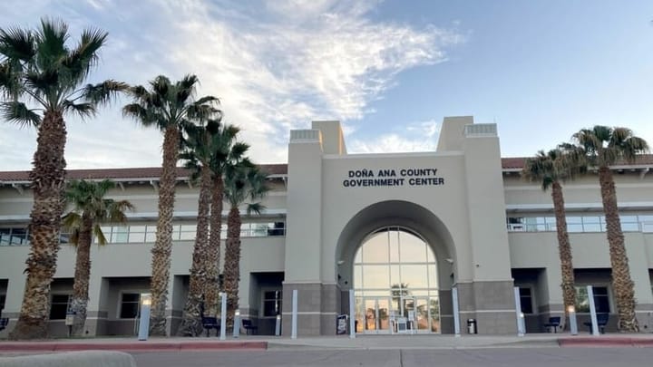 Exterior of the Doña Ana County Government Center in Las Cruces showing its arched glass entrance, beige facade and rows of palm trees under a partly cloudy sky.