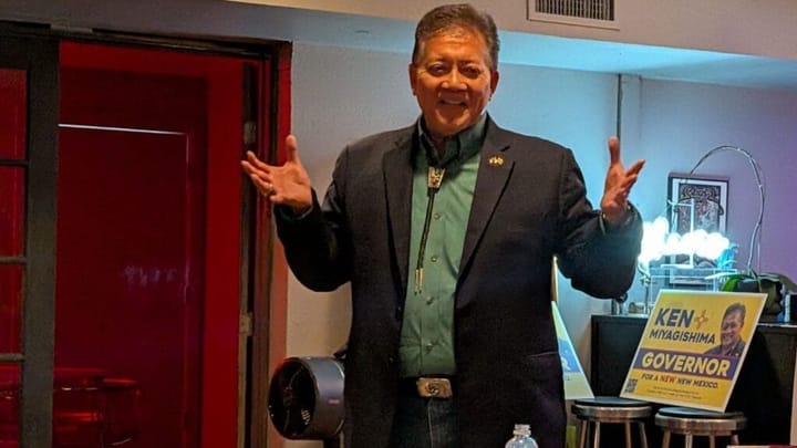 Former Las Cruces Mayor Ken Miyagishima speaks to supporters indoors, standing with his arms raised near campaign signs during his run for New Mexico governor.