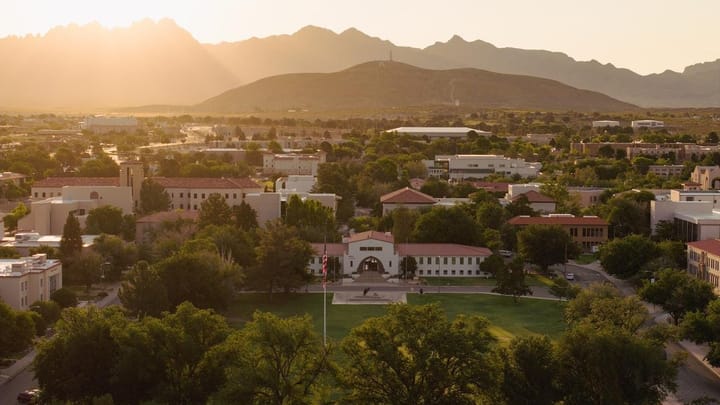 A wide aerial view of the New Mexico State University campus at sunset, with campus buildings, trees and the Organ Mountains in the background glowing in warm light.