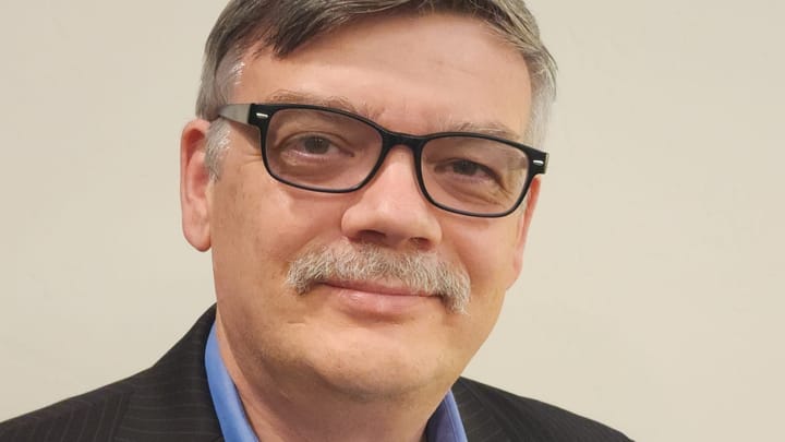 Head-and-shoulders portrait of Sergey Dryga wearing black-framed glasses, a blue collared shirt and a dark pinstriped blazer against a plain light background.
