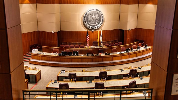 The New Mexico State Senate chamber in Santa Fe, showing rows of empty desks, maroon chairs, and the state seal mounted on the wall behind the podium.
