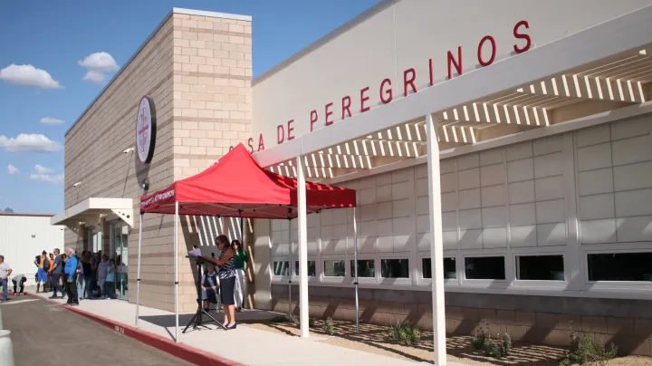 Exterior of Casa de Peregrinos in Las Cruces, with people lined up outside the food pantry and a small outdoor gathering under a red canopy near the entrance.