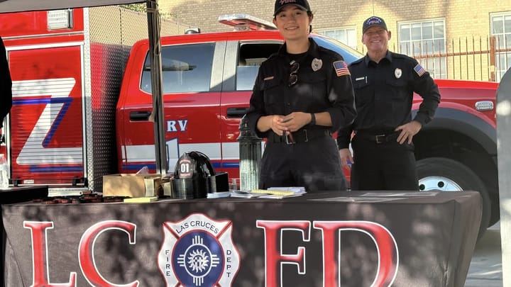 Two Las Cruces Fire Department firefighters stand behind an LCFD information table, with a fire engine parked behind them at an outdoor community event.