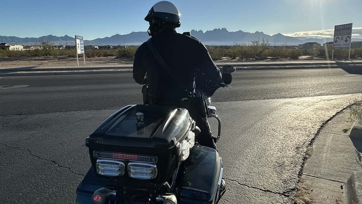 A Las Cruces police officer on a motorcycle watches traffic on Red Hawk Golf Road with the Organ Mountains visible in the distance.