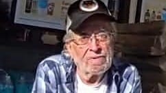 A 68-year-old man wearing a dark baseball cap, glasses, and a blue plaid shirt sits indoors with his arms crossed. He has gray facial hair and a serious expression.