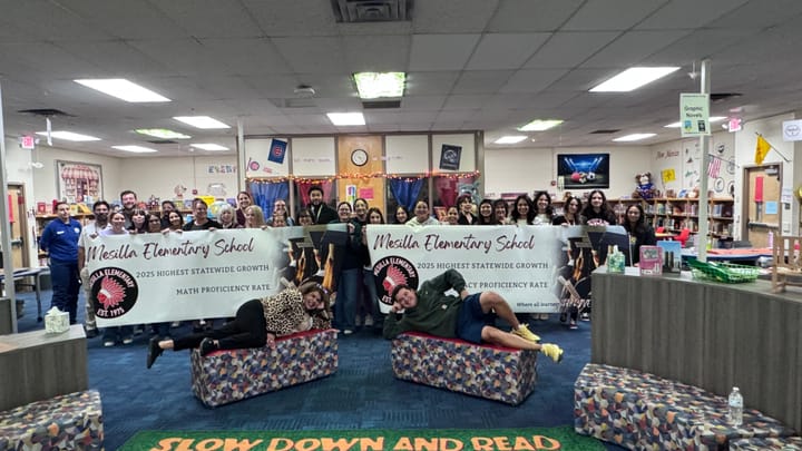 A large group of Mesilla Elementary staff pose in the school library with two banners celebrating statewide growth in literacy and math, with two staff members playfully posing in front.