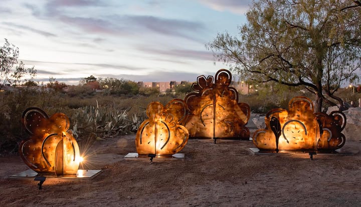 Metal sculpture panels shaped like desert plants glow from internal lighting at dusk in a landscaped Las Cruces park, with trees, cacti and buildings visible in the background.