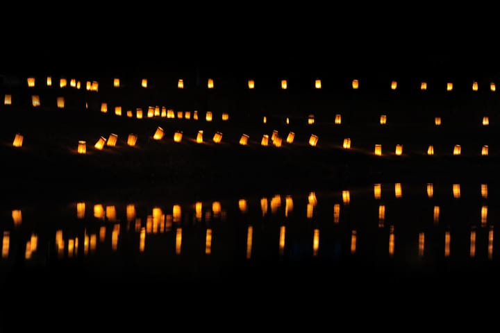 Rows of glowing luminarias line a dark pathway at night, their warm light reflecting off the water below.