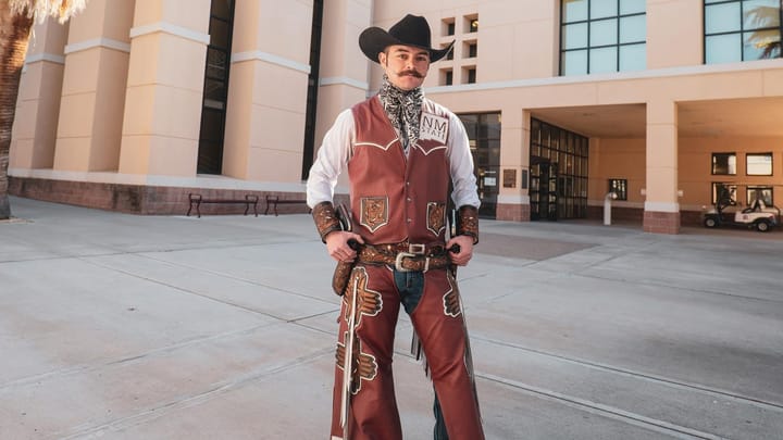 Pistol Pete, the New Mexico State University Aggies mascot, stands outside an NMSU campus building wearing a maroon cowboy outfit and hat.