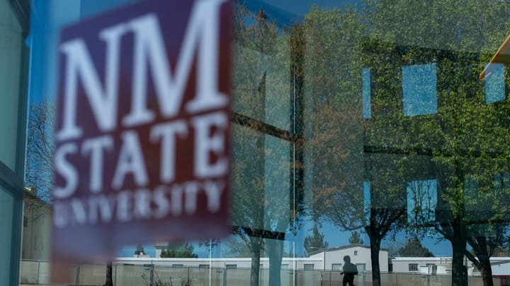 A window reflects trees and campus buildings near New Mexico State University, with the NMSU logo visible on the glass.