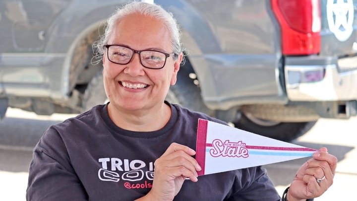 A woman wearing a TRIO program T-shirt smiles while holding a New Mexico State University pennant during an outreach event focused on college readiness.