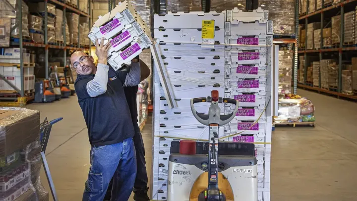 A worker lifts stacked produce boxes onto his shoulder inside a warehouse, standing next to a pallet jack loaded with shrink-wrapped cartons, with shelves of food visible in the background.