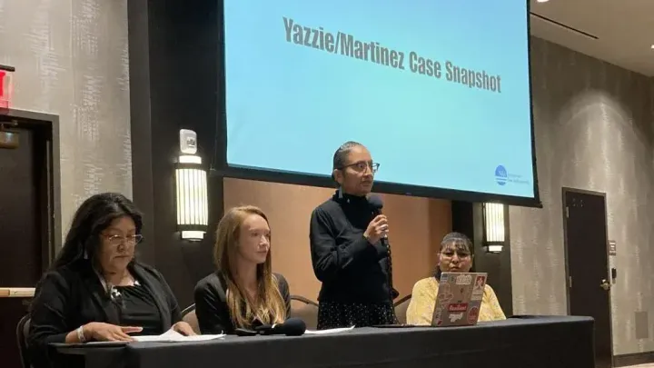 Four speakers sit at a panel table as a presenter stands with a microphone in front of a screen titled Yazzie/Martínez Case Snapshot during a public meeting.
