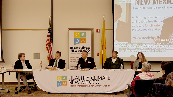 Ken Miyagishima sits at a panel table during a Healthy Climate New Mexico town hall, with other panelists, audience members and presentation slides visible in a meeting room.