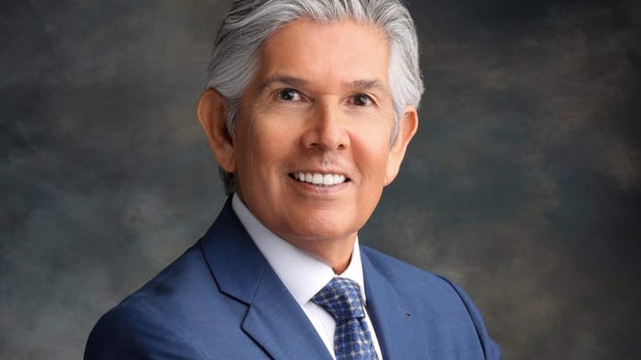 Duke Martinez, with gray hair wearing a blue suit and patterned tie, poses for a studio portrait with his arms crossed.