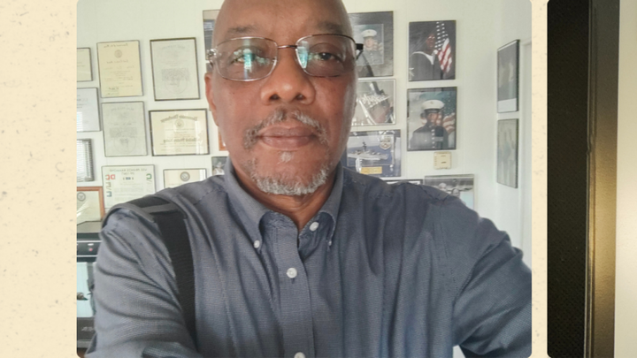 Volunteer ombudsman Kevin Thompson stands indoors wearing glasses and a button-down shirt, with framed certificates and military photos displayed on the wall behind him.