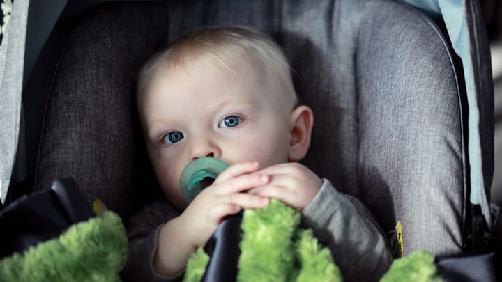 A baby with blond hair sits in a gray car seat holding a green blanket and a pacifier while looking toward the camera.