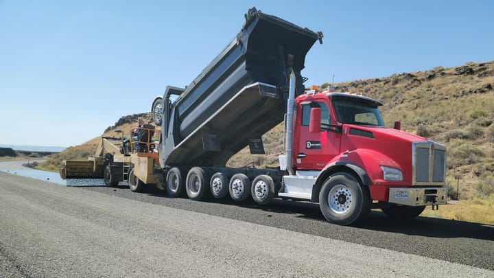 A dump truck unloads asphalt as road construction equipment repaves a highway in a rural area.