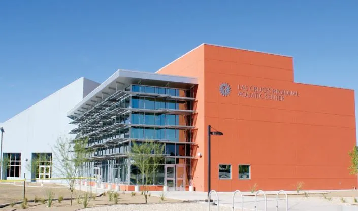 Exterior view of the Las Cruces Regional Aquatic Center, a modern building with an orange facade, large glass windows, and metal sunshades under a clear blue sky.