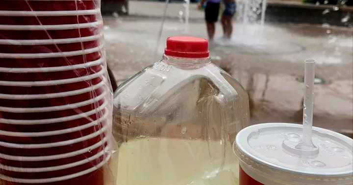 Raising Cane’s cups and lemonade jug at a previous Lunchbox Lemonade event on Plaza de Las Cruces, with children playing in the splash pad behind