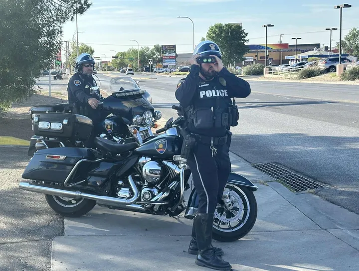 Two Las Cruces police officers on motorcycles conduct a traffic enforcement operation, with one officer using binoculars to monitor vehicles along El Paseo Road