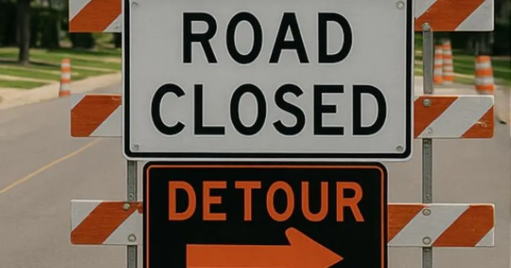 Portable road sign reading “Road Closed” above a black-and-orange “Detour” sign with a right-pointing arrow, set up on a street with orange traffic barrels in the background