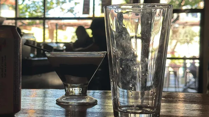 A small martini-style glass with a dark layered drink sits on a wooden table beside an empty pint glass, with sunlight streaming through large windows in the background.