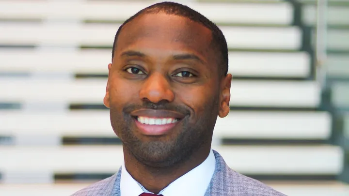 Joe Fields, New Mexico State University’s newly named athletics director, smiles while wearing a light gray suit and red tie in front of blurred steps