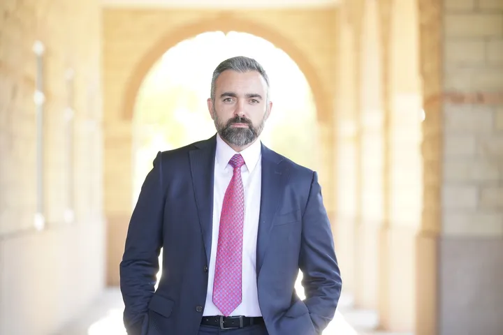 New Mexico Attorney General Raúl Torrez stands outdoors under an arched walkway, wearing a navy suit, white shirt, and red patterned tie, looking directly at the camera.