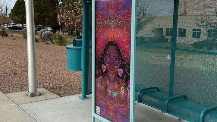 A colorful portrait artwork displayed on a teal bus shelter in Las Cruces for the Art Stop program. The piece shows a woman surrounded by hearts, flowers and vivid patterns along Route 2.