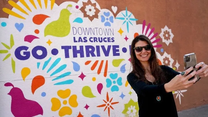 A woman takes a selfie in front of a colorful mural in downtown Las Cruces that reads “Downtown Las Cruces Go Thrive.”