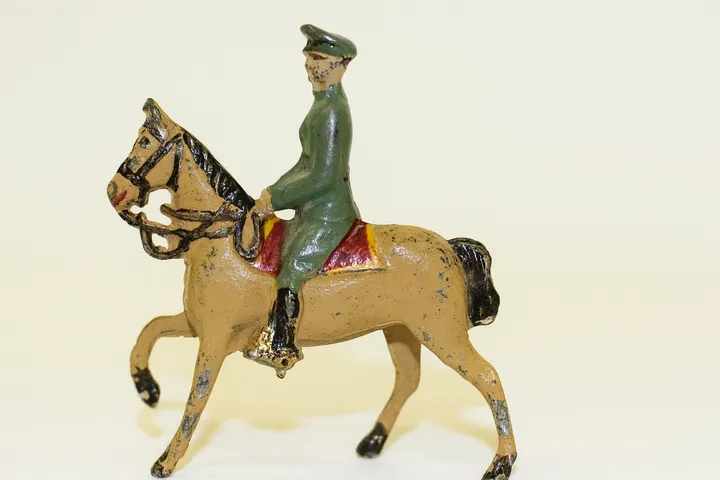ChatGPT said:  A vintage cast-metal toy soldier figurine in a green uniform and cap rides a tan horse with a red saddle, shown against a plain light background.