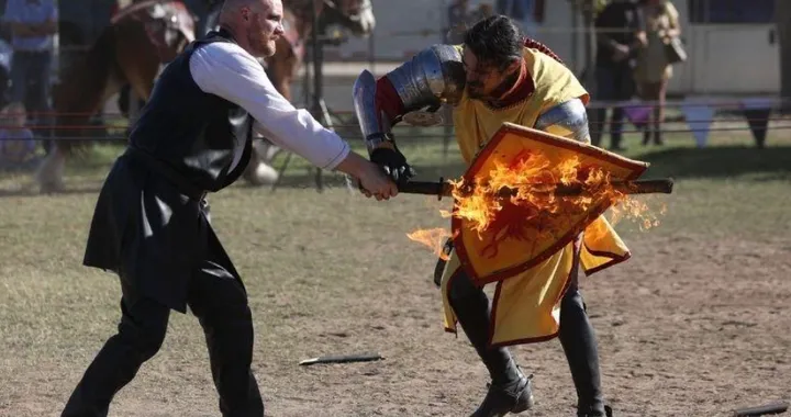 Two armored performers stage a combat scene at a Renaissance fair, with one holding a flaming shield while the other strikes it with a weapon.