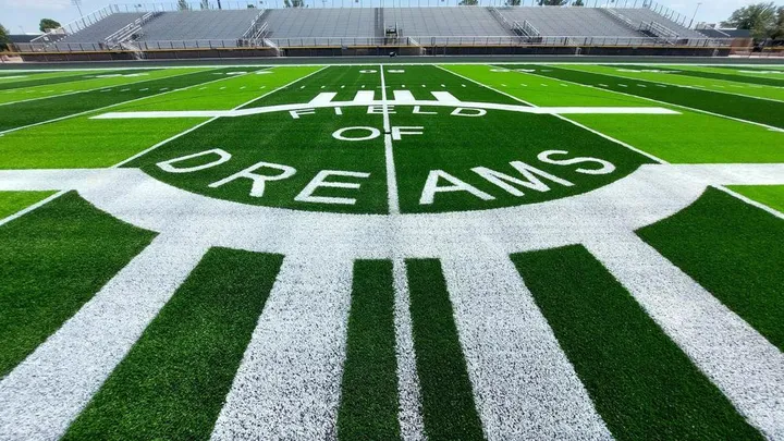 White letters spelling “Field of Dreams” are painted on the bright green turf at the Las Cruces Public Schools football stadium, with empty bleachers visible in the background.