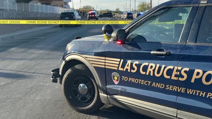 Las Cruces Police Department vehicle blocks a street behind yellow crime scene tape as officers investigate in the distance on a sunny afternoon.